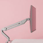 Um suporte de monitor articulado, de cor prateada, segura um monitor de tela plana. O fundo é de uma cor rosa pastel sólida.
