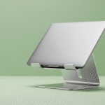 Um suporte de notebook minimalista, em alumínio escovado, a segurar um portátil aberto. O fundo é de uma cor verde pastel suave e sólida.